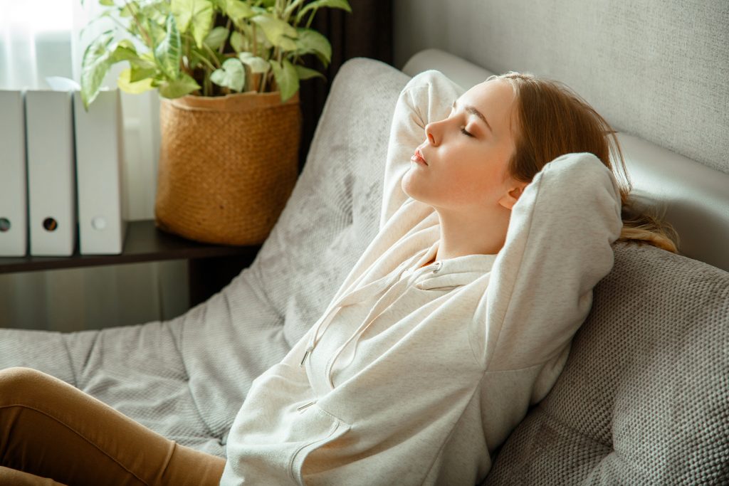 Happy calm young woman relax sitting on comfort couch at home interior. Teen girl resting, bliss enjoy. Portrait of woman resting with closed eyes